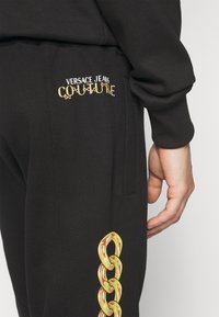 Černé tepláky s žlutou vyšívanou značkou "VERSACE JEANS COUTURE" a zlatým řetězcem na boku. Měkká textilie.