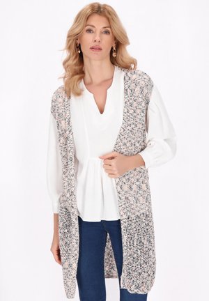 Blonde vrouw die een witte blouse, blauwe spijkerbroek en een lange gebreide mouwloze cardigan met een roze en grijs patroon draagt.