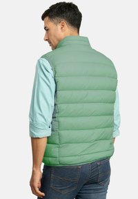Chaleco acolchado verde claro con secciones de acolchado horizontal. Presenta un cuello alto y se lleva sobre una camisa azul claro, combinado con unos vaqueros oscuros.