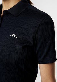 Marinblå polo skjorta med texturerat vågmönster, korta ärmar och krage. Har en vit logotyp på vänster bröst.