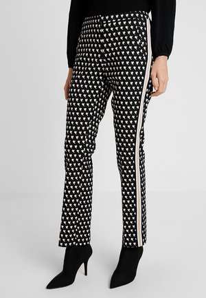 Pantalon noir avec motifs de cœurs blancs, bandes latérales blanches, porté avec des bottines noires à talons pointus et un haut noir à manches longues.