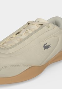 Baskets en cuir beige avec des lacets blancs, une semelle marron et un petit logo crocodile foncé sur le côté.