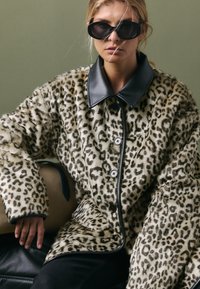 Cappotto con stampa leopardata e texture in pelliccia sintetica, colletto in pelle nera e grandi bottoni. Le maniche sono oversize, offrendo una vestibilità comoda.