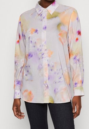 Chemise à manches longues boutonnée avec des motifs floraux pastel doux en orange, violet et vert sur fond clair, portée avec un pantalon foncé.