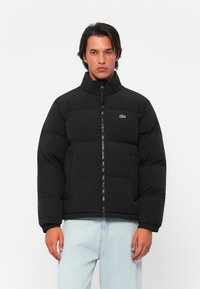 Veste matelassée noire avec col montant, fermeture éclair et poignets côtelés. Présente un logo vert sur la poitrine gauche. Portée sur un jean bleu clair.