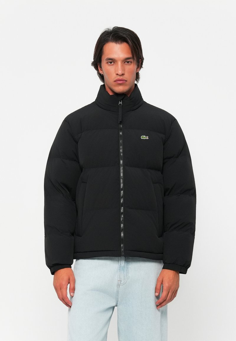 Veste matelassée noire avec col montant, fermeture éclair et poignets côtelés. Présente un logo vert sur la poitrine gauche. Portée sur un jean bleu clair.