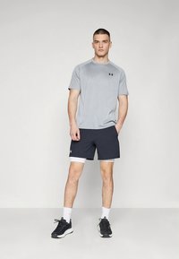 Grijs atletisch t-shirt met zwart logo, gecombineerd met marineblauwe shorts met witte accenten. Model draagt zwarte sneakers met witte zolen en witte sokken.