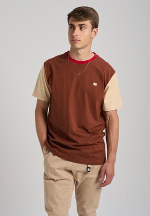 Junger Mann trägt ein braunes und beiges T-Shirt mit rotem Kragen und beigen Hosen, steht mit einer Hand in der Tasche und schaut zur Seite.