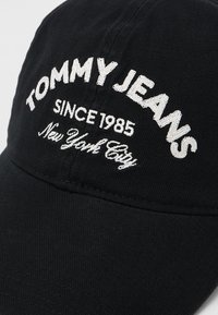 Svart baseballcap med hvit brodert tekst som sier "TOMMY JEANS SINCE 1985 New York City" på frontpanelet.