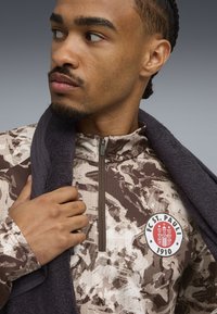 Brun og beige camouflage zip-up jakke med logo-patch, der har en høj krave og struktureret stof. Håndklæde draperet over skulderen.