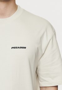 Ljusbeige kortärmad T-shirt i bomull, med en svart "PEGADOR"-logotyp på vänster bröstområde. Slät textur, rundad halsdesign.