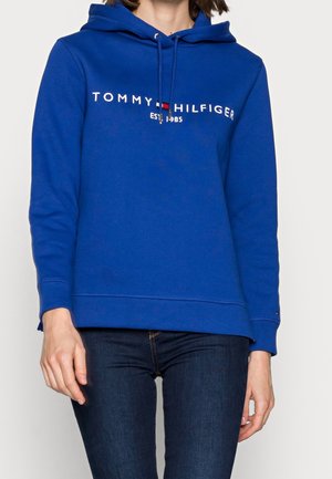 Kvinde iført en blå Tommy Hilfiger hættetrøje med logo og teksten "EST 1985", kombineret med mørkeblå jeans, stående foran en ensfarvet baggrund.
