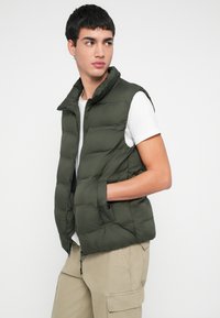 Mladý muž v tmavozelenej vatovanej veste cez biele tričko a béžové nohavice s bočnými vreckami, stojaci s jednou rukou v vrecku, hľadí do strany.