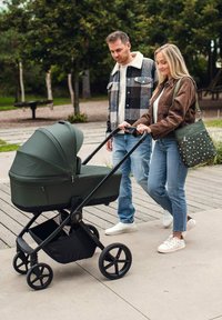 Zelený kočárek s měkkou látkovou stříškou, černým rámem a velkými koly. Dospělí kráčejí s puntíkovanou taškou, dřevěným a travnatým pozadím.