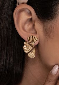 Primer plano de un pendiente en forma de mariposa dorada llevado en la oreja izquierda, con cabello oscuro y una mano con esmalte de uñas rosa pálido cerca de la barbilla.