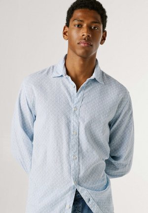 Noor meeshall lühikeste lokkis juustega ja helesinise mustrilise nööbitava särgiga, seistes ühevärvilise valge tausta ees.