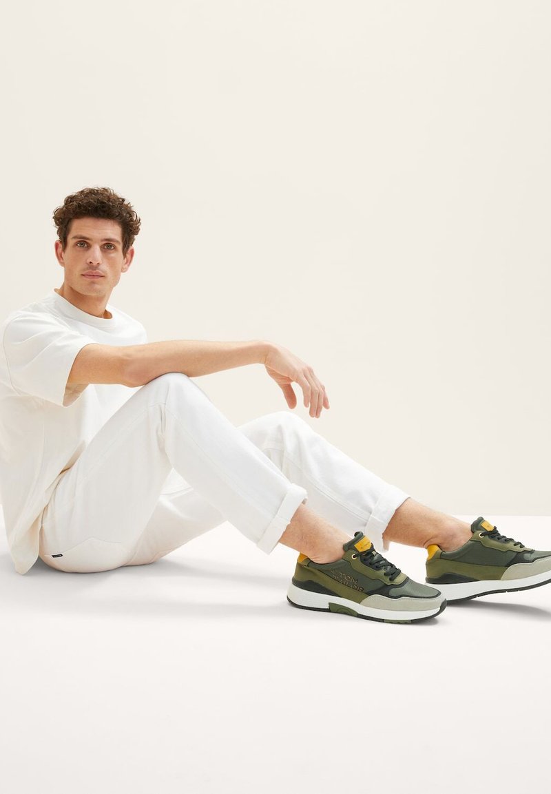 Sneaker multicolori con un mix di verde oliva, grigio e accenti gialli, abbinati a una maglietta bianca casual e jeans bianchi.