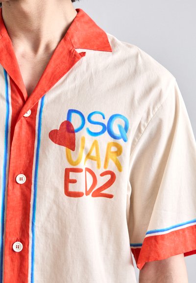 Chemise à manches courtes en coton beige avec col et poignets rouges. Présente un graphisme textuel multicolore indiquant "DSQ♥VAR ED2" avec des accents bleus et jaunes.