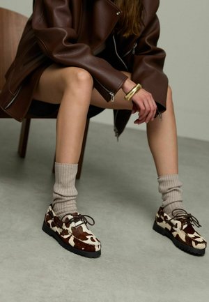 Braun-weiße Cowprint-Schuhe aus Wildleder mit einer Schnürdesign und einer robusten schwarzen Sohle, kombiniert mit gerippten beigen Socken.