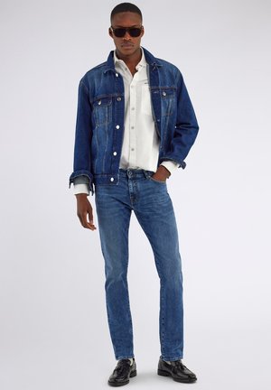 Hombre con gafas de sol oscuras, camisa blanca, chaqueta vaquera azul, vaqueros ajustados azules y zapatos de cuero negros, de pie con una mano en el bolsillo.