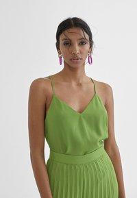 Groene mouwloze top met een V-hals, gemaakt van gladde stof, gedragen met een bijpassende geplooide rok. Opvallende roze ringoorbellen complementeren de outfit.