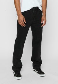 Jeans negri din denim cu croială dreaptă, având cinci buzunare, bucle pentru curea și închidere cu nasture. Poartă pantofi sport negri, din material texturat.