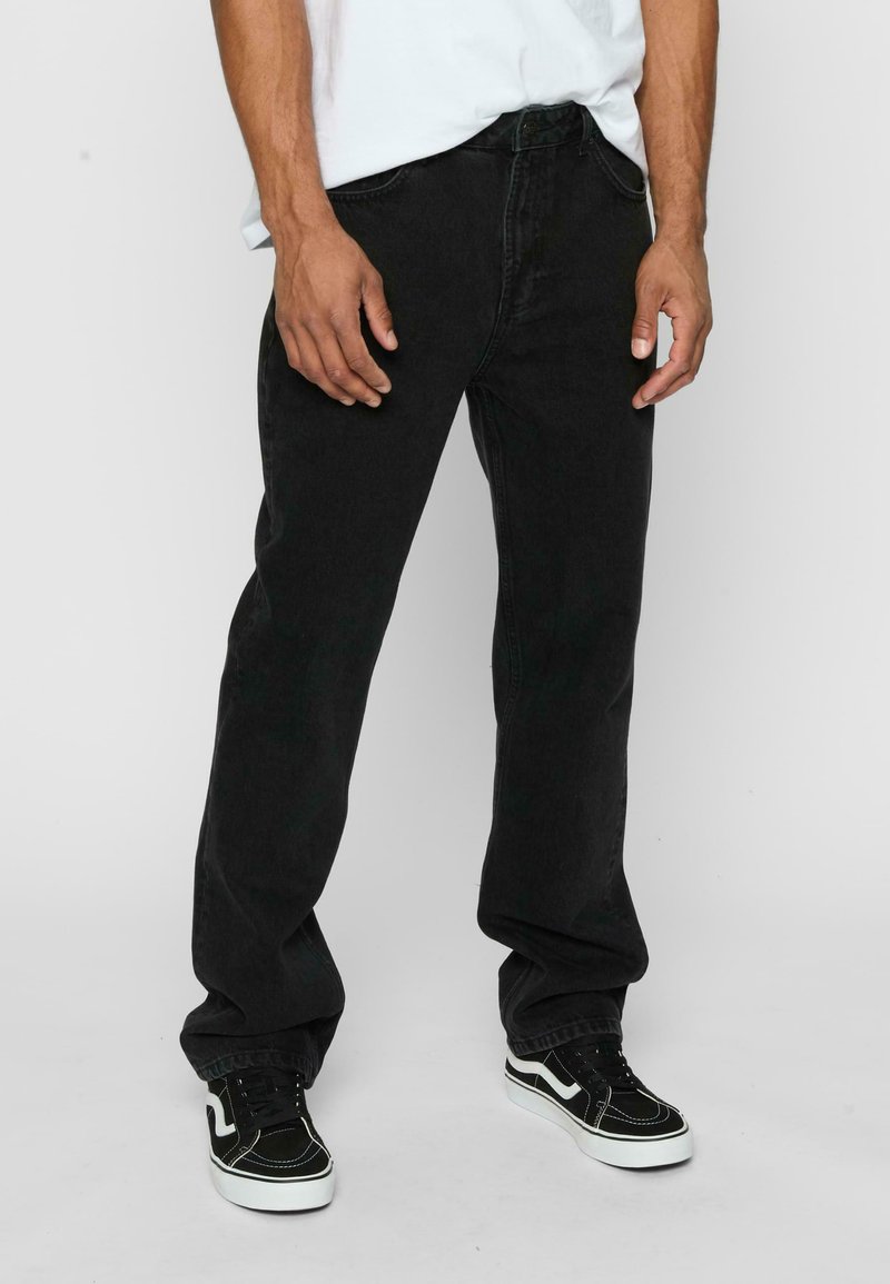 Jeans negri din denim cu croială dreaptă, având cinci buzunare, bucle pentru curea și închidere cu nasture. Poartă pantofi sport negri, din material texturat.