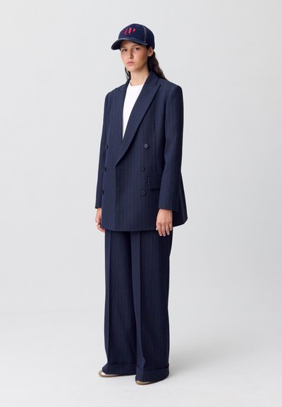 Blazer de doble botonadura a rayas marino con pantalones de pierna ancha a juego, acompañado de una gorra azul marino con un diseño en rojo.