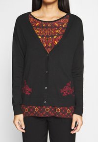 Schwarze Strickjacke mit V-Ausschnitt, Knöpfen auf der Vorderseite und roter Blumenstickerei auf den Taschen. Darunter ein rotopatterniertes Oberteil mit gezacktem Saum.
