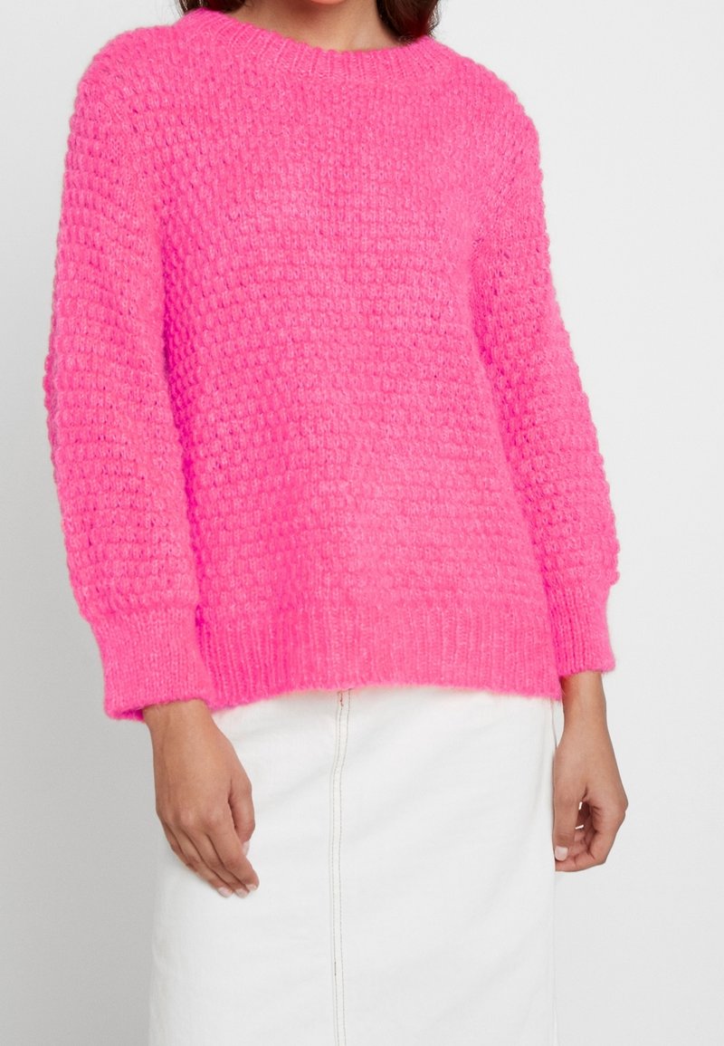 Pull en laine flou de couleur rose vif avec un design tricoté texturé, col rond et poignets côtelés, porté sur une jupe blanche.