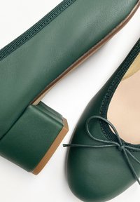 Des ballerines en cuir vert avec un talon carré, un bout rond et un nœud décoratif. Un bord en élastique texturé souligne le design.