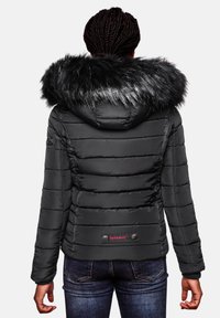 Black puffer jacket with a detachable faux fur hood, quilted texture, and red logo detail at the waist, paired with dark jeans.
