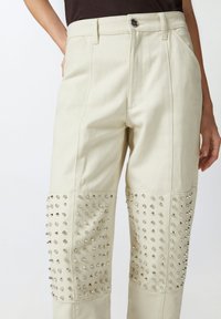 Pantalone kremaste boje od traper materijala, s podebljanim detaljima na donjem dijelu nogu i klasičnim dizajnom s pet džepova.