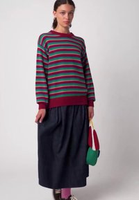 Gestreifter Pullover in Grün-, Rot- und Dunkellila-Tönen; kombiniert mit einem schwarzen Plisseerock. Das Model hält eine mehrfarbige Tasche.