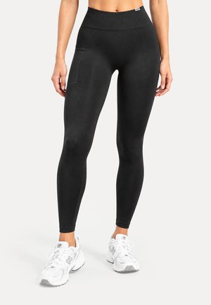 Femme portant un legging noir taille haute avec une poche latérale et des chaussures de sport blanches, debout devant un fond uni.