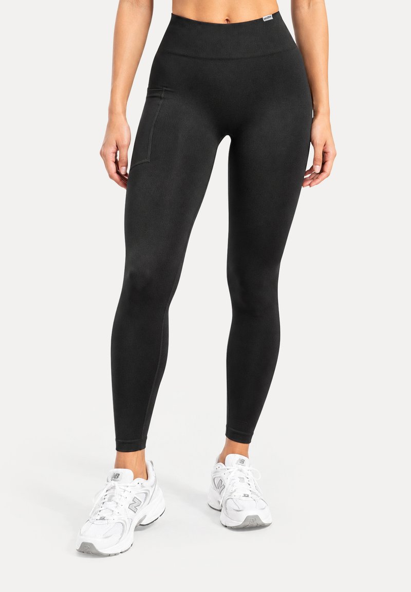 Femme portant un legging noir taille haute avec une poche latérale et des chaussures de sport blanches, debout devant un fond uni.