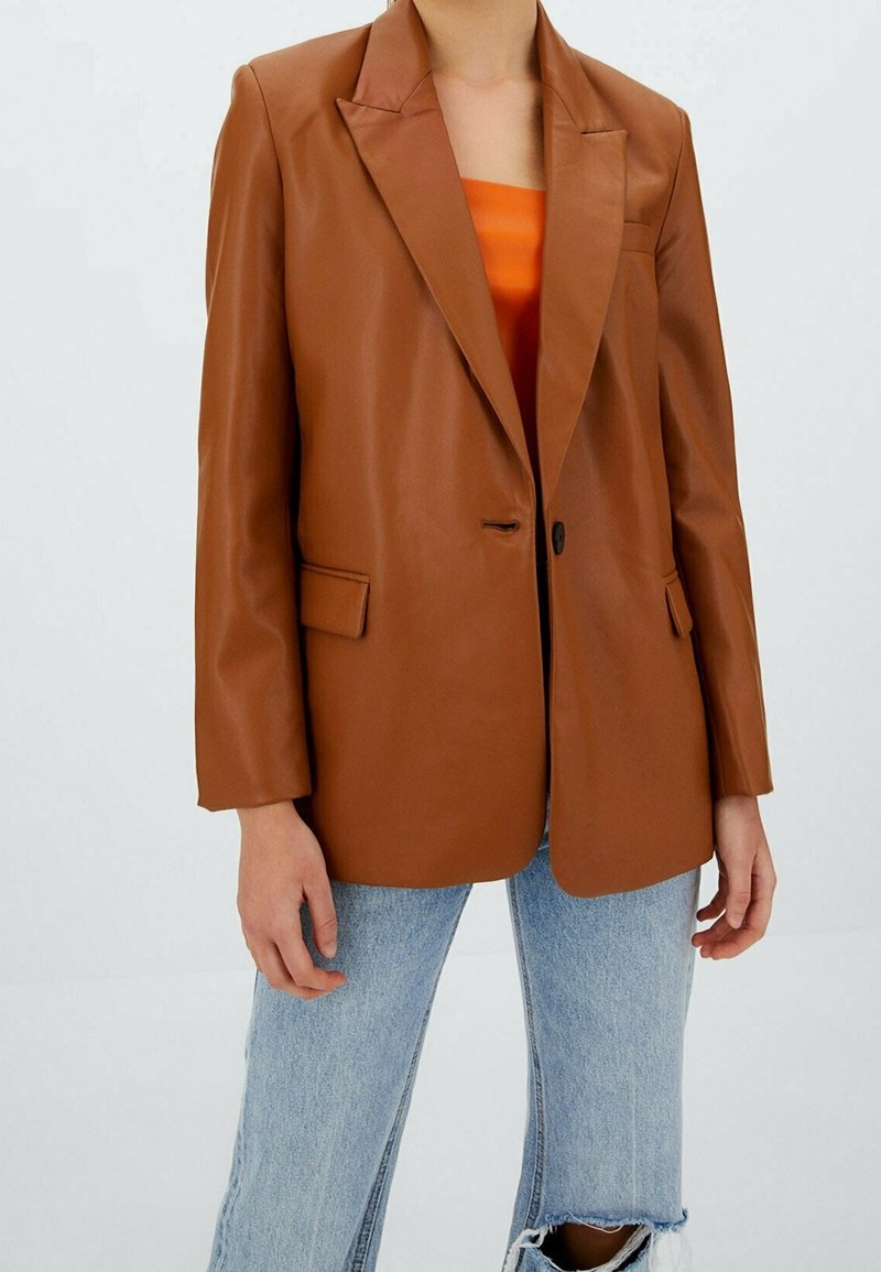 Bruine leren blazer met een enkele knoop, notched revers, twee voorzakken en lange mouwen, gecombineerd met een feloranje top en versleten spijkerbroek.