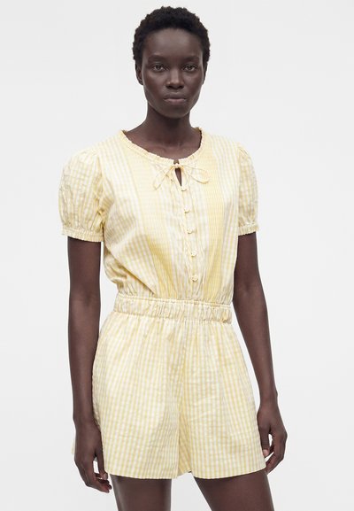 Γυναίκα που φορά κοντό ολόσωμο ρούχο με κίτρινες και λευκές ρίγες, κουμπιά, ελαστική μέση και δεμένο ντεκολτέ σε μονόχρωμο φόντο.