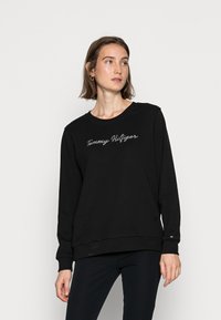 Felpa nera con maniche lunghe, colletto arrotondato, con la scritta "Tommy Hilfiger" ricamata in corsivo sul davanti. Tessuto morbido, vestibilità comoda.