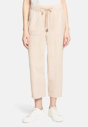 Femme portant un pantalon beige court avec une taille élastique et un cordon de serrage, associé à des baskets blanches sur un fond uni.