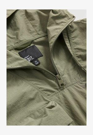 Jachetă ușoară de culoare verde măsliniu, cu închidere prin fermoar pe jumătate, etichetă din material și textură netedă. Designul include o glugă și cusături laterale.