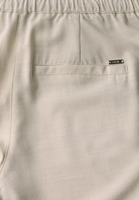 Ljust beige, texturerat tyg på byxorna har en bakficka med en liten metallisk logotag, vilket visar en avslappnad och stilren design.