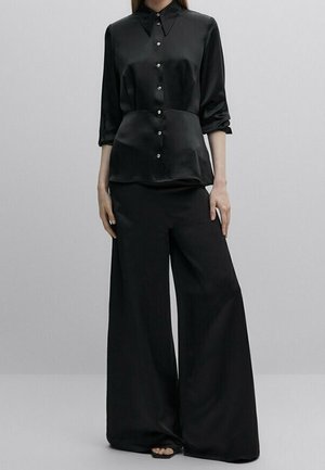 Femme portant une chemise noire en satin à boutons avec manches trois quarts et pantalon noir à jambes larges sur un fond gris uni.
