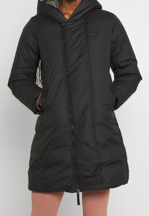 Manteau matelassé noir à capuche avec fermeture éclair frontale porté sur un pantalon beige côtelé, mains dans les poches, vue recadrée du menton à mi-cuisse.