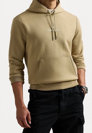 Sweat à capuche beige avec poche avant et cordons, porté avec un pantalon cargo noir et une montre au poignet gauche.