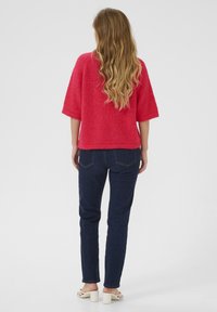 Pull en tricot fluo rose avec de larges manches, associé à un jean en denim bleu foncé et des sandales à talons carrés blanches. Vue arrière.