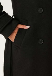 Cappotto di lana nera con una texture liscia, dotato di chiusura a bottoni e tasca laterale. Primo piano di una mano appoggiata nella tasca.