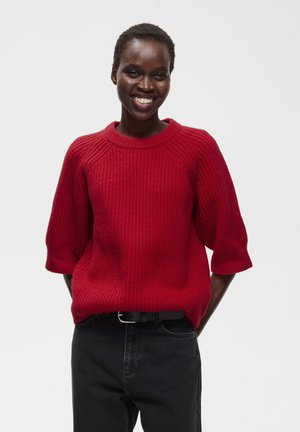 Personne souriante portant un pull rouge côtelé à manches trois-quarts et un pantalon noir avec une ceinture noire sur un fond uni.