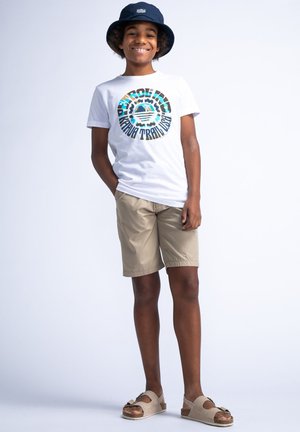 Camiseta blanca con gráfico circular colorido, pantalones cortos caqui y sandalias beige. Los accesorios incluyen un sombrero de cubo azul marino. Atuendo casual.