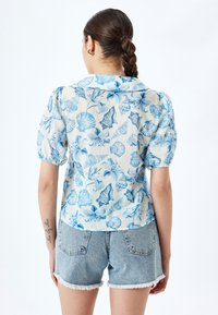 Kariertes Hemd aus leichtem Stoff mit einem blauen Blumen- und Muschel-Muster auf hellem Hintergrund, mit Puffärmeln und Kragen.